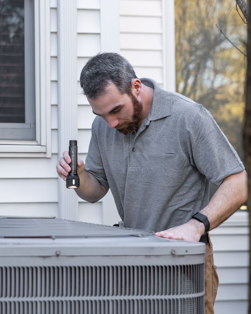 A technician inspects a residential air conditioning unit with a flashlight.